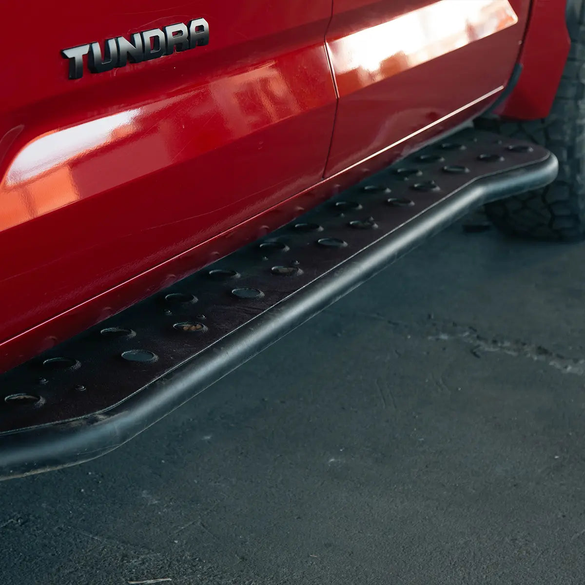 A close-up of a black metal truck side step bar mounted below a red pickup truck, with the TUNDRA badge visible and pavement underneath.