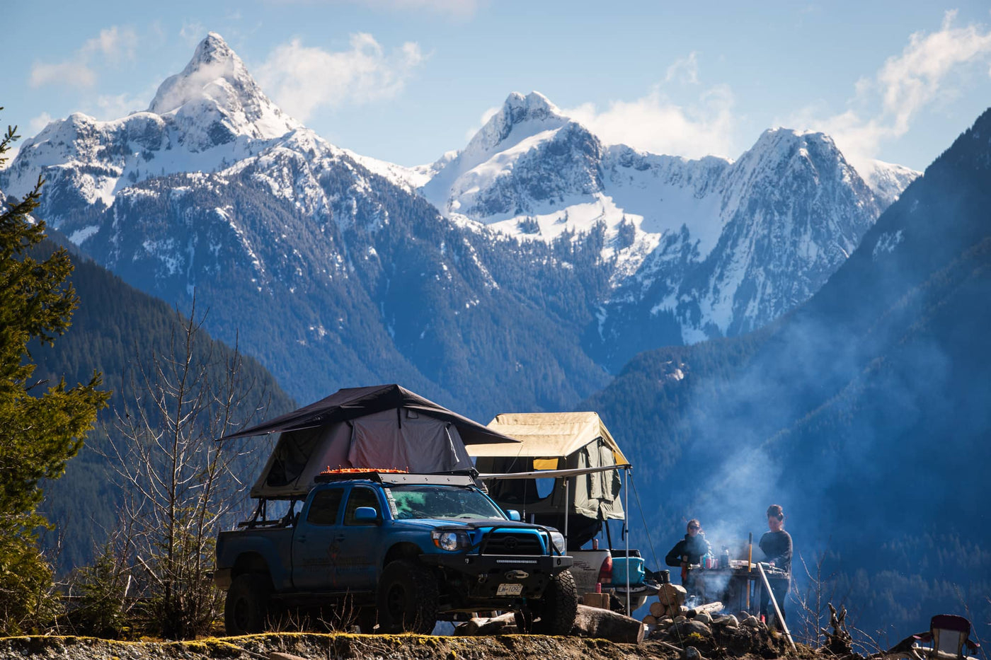 Two trucks with rooftop tents parked in a mountainous area with snow-capped peaks.