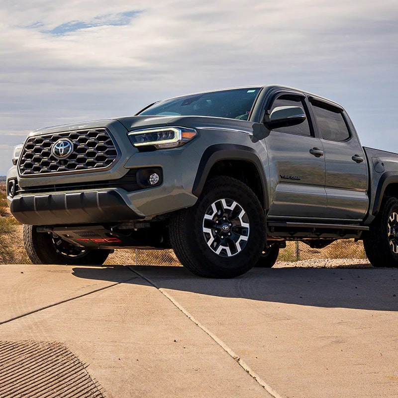 Gray Tacoma truck parked on a concrete surface with a cloudy sky in the background with white fog lights