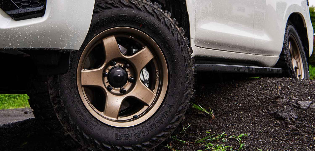 Close-up of a vehicle tire with a bronze alloy wheel on a dirt road.
