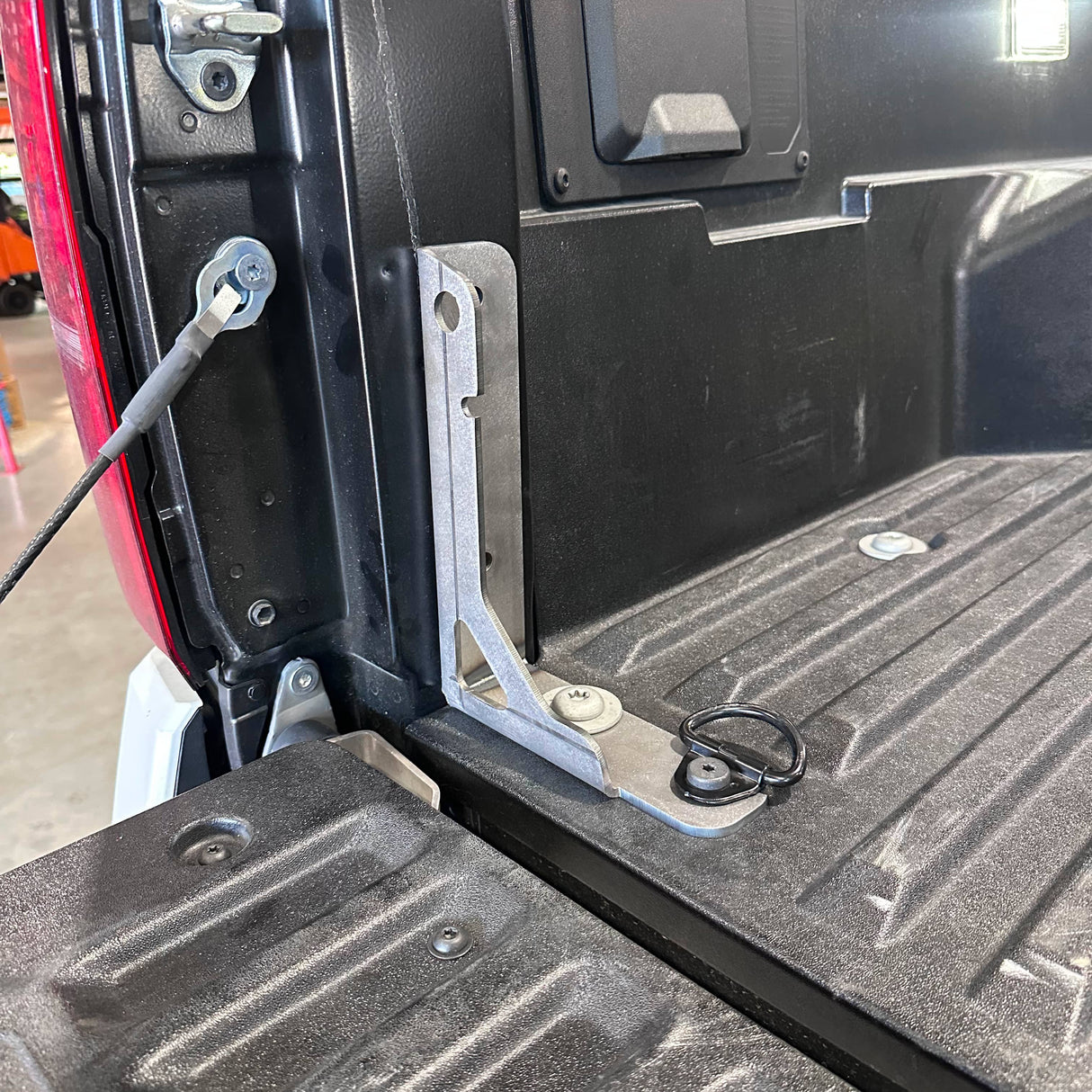 A close-up of a black metal truck bed accessory bracket mounted on the side of a truck bed, with a textured black liner floor and tailgate visible.