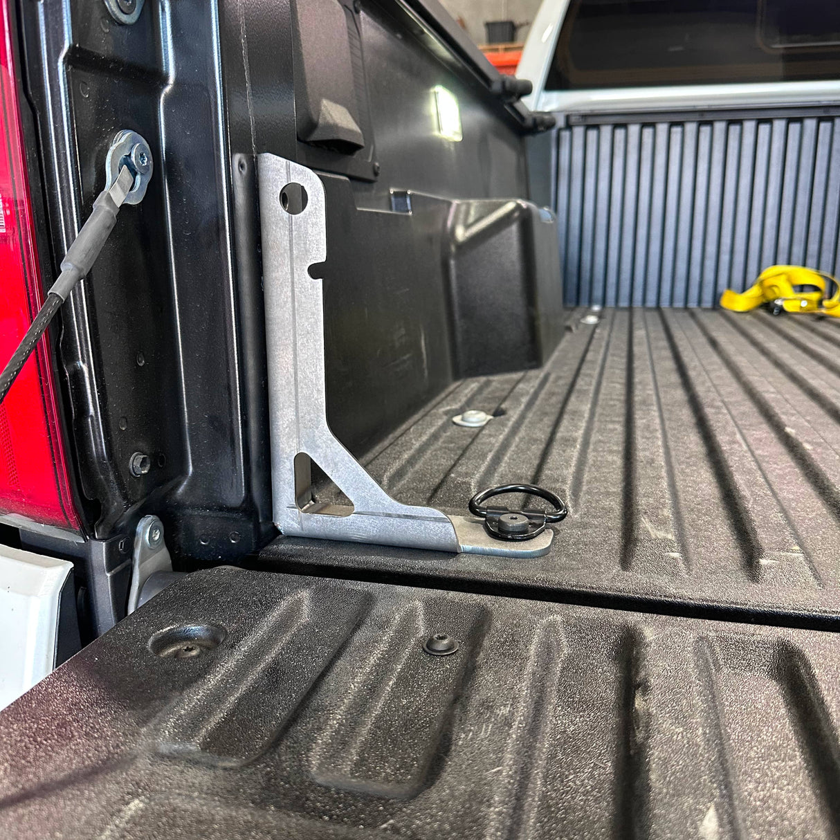 A silver metal truck bed ladder bracket installed on the side of a black pickup truck bed, with a black textured truck bed liner and a yellow strap visible in the background.