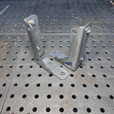 Two galvanized steel angle brackets with mounting plates, standing upright on a perforated metal workbench surface.