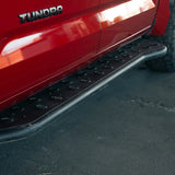 A close-up of a black metal truck side step bar mounted below a red pickup truck, with the TUNDRA badge visible and pavement underneath.