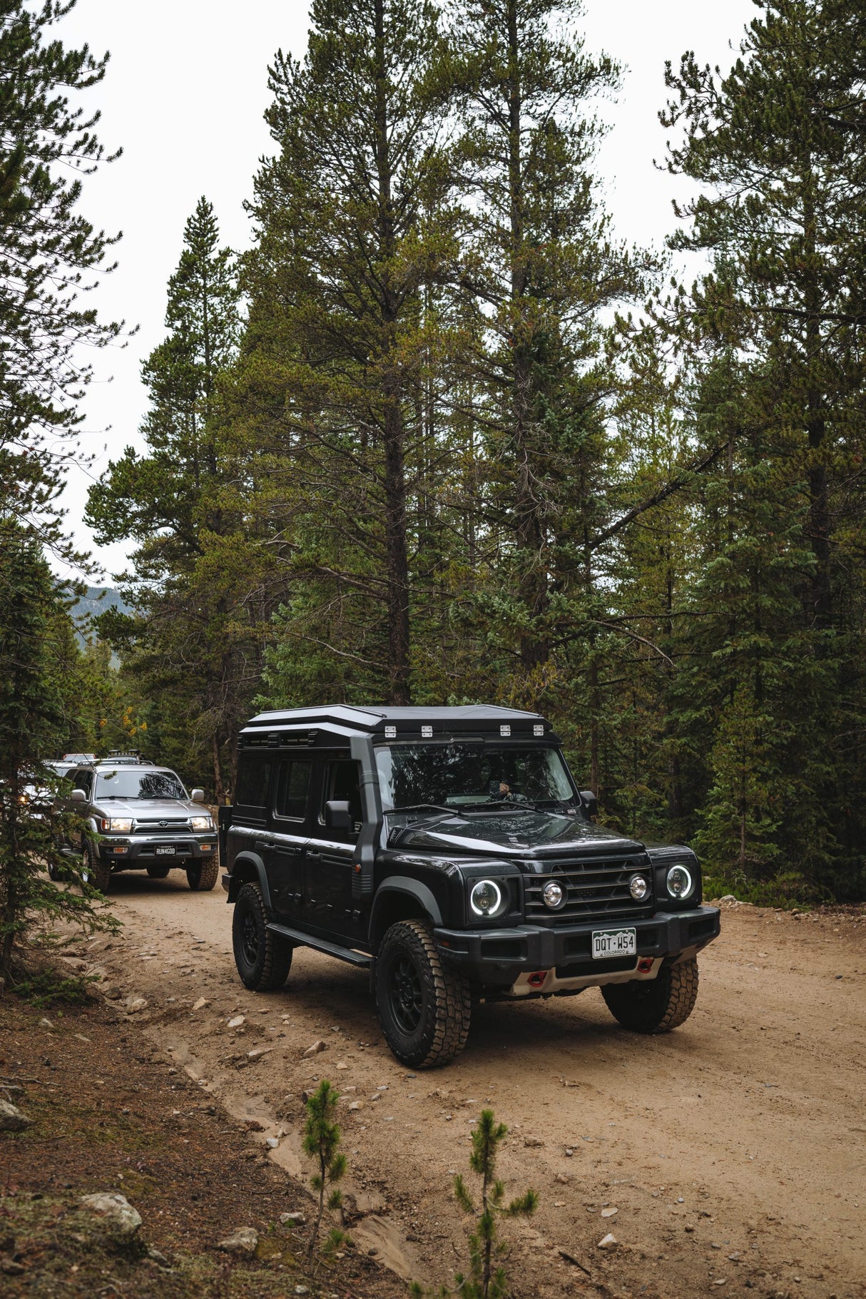 A black off-road SUV with a roof rack and front bumper light is driving on a dirt trail surrounded by tall pine trees. Another vehicle is visible in the background.