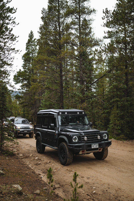 A black off-road SUV with a roof rack and front bumper light is driving on a dirt trail surrounded by tall pine trees. Another vehicle is visible in the background.