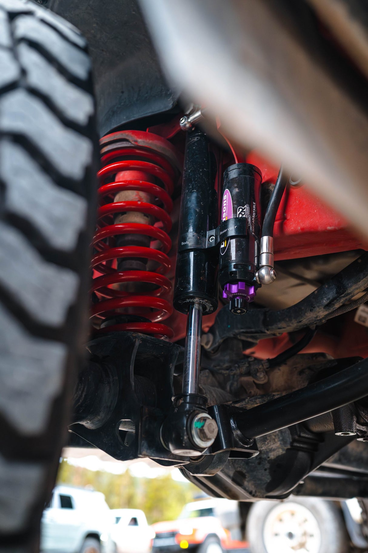 Close-up of a vehicle's undercarriage showing a red coil spring, black shock absorber, and suspension components, photographed outdoors with blurred vehicles in the background.