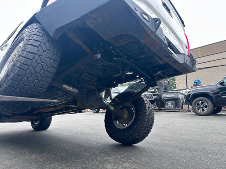 Close-up low-angle view of the undercarriage and rear suspension of a lifted black pickup truck, showing the rear tire, exhaust area, and mechanical components in an outdoor lot.