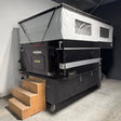A black and silver rooftop camper tent with a wooden three-step stair, photographed in a warehouse or garage setting.