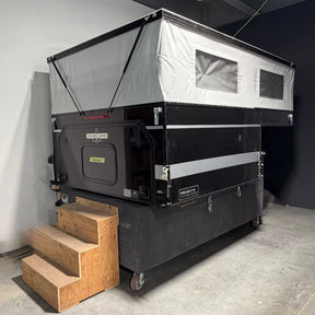 A black and silver rooftop camper tent with a wooden three-step stair, photographed in a warehouse or garage setting.