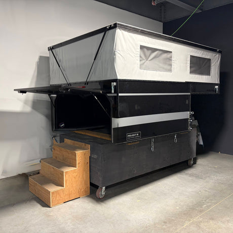 A black and white rooftop camper trailer with an attached pop-up tent, shown in a garage setting with wooden stairs in the foreground.