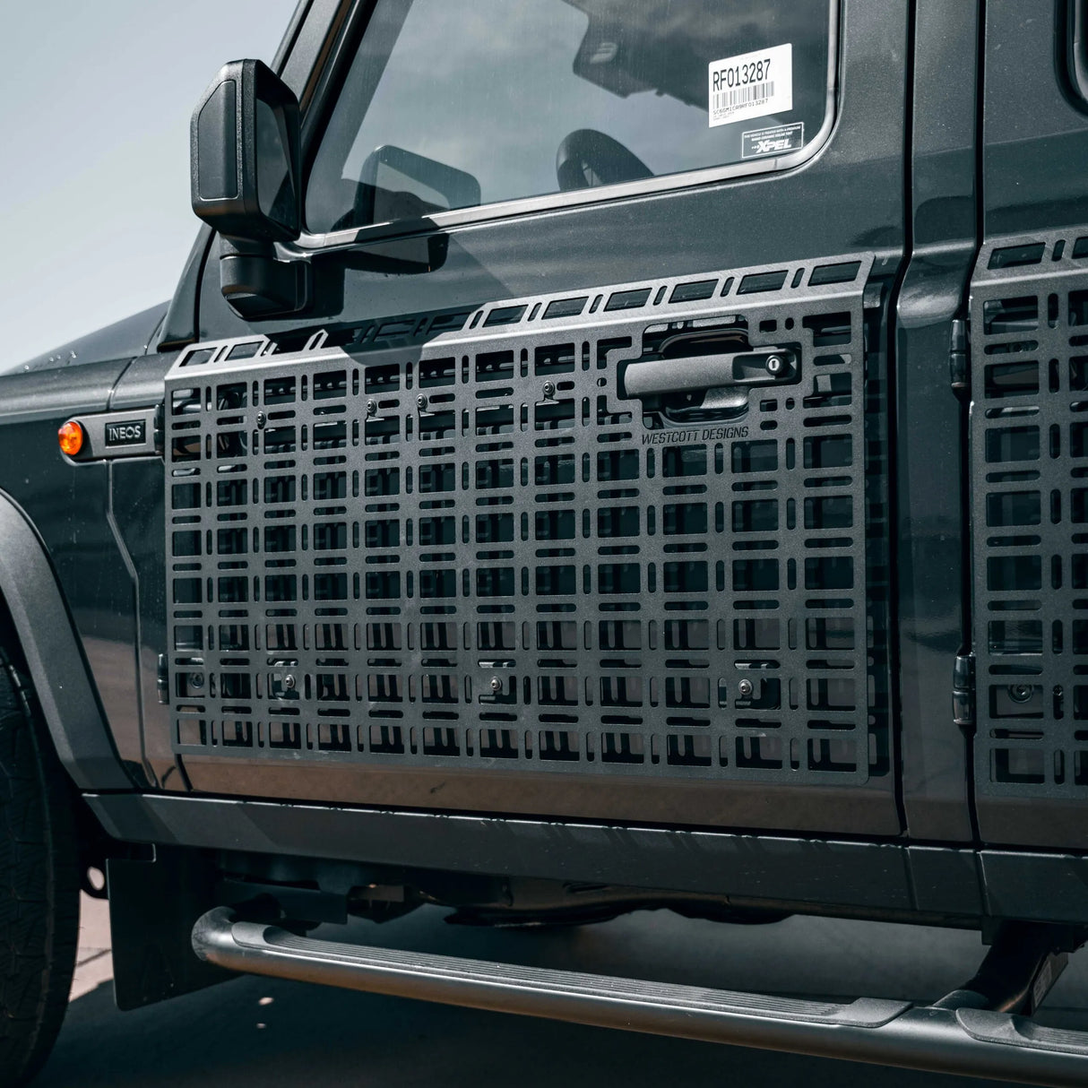 Black metal side step grates installed on the side of a dark gray off-road vehicle, featuring a grid pattern and a central handle cutout.