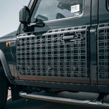 Black metal side step grates installed on the side of a dark gray off-road vehicle, featuring a grid pattern and a central handle cutout.