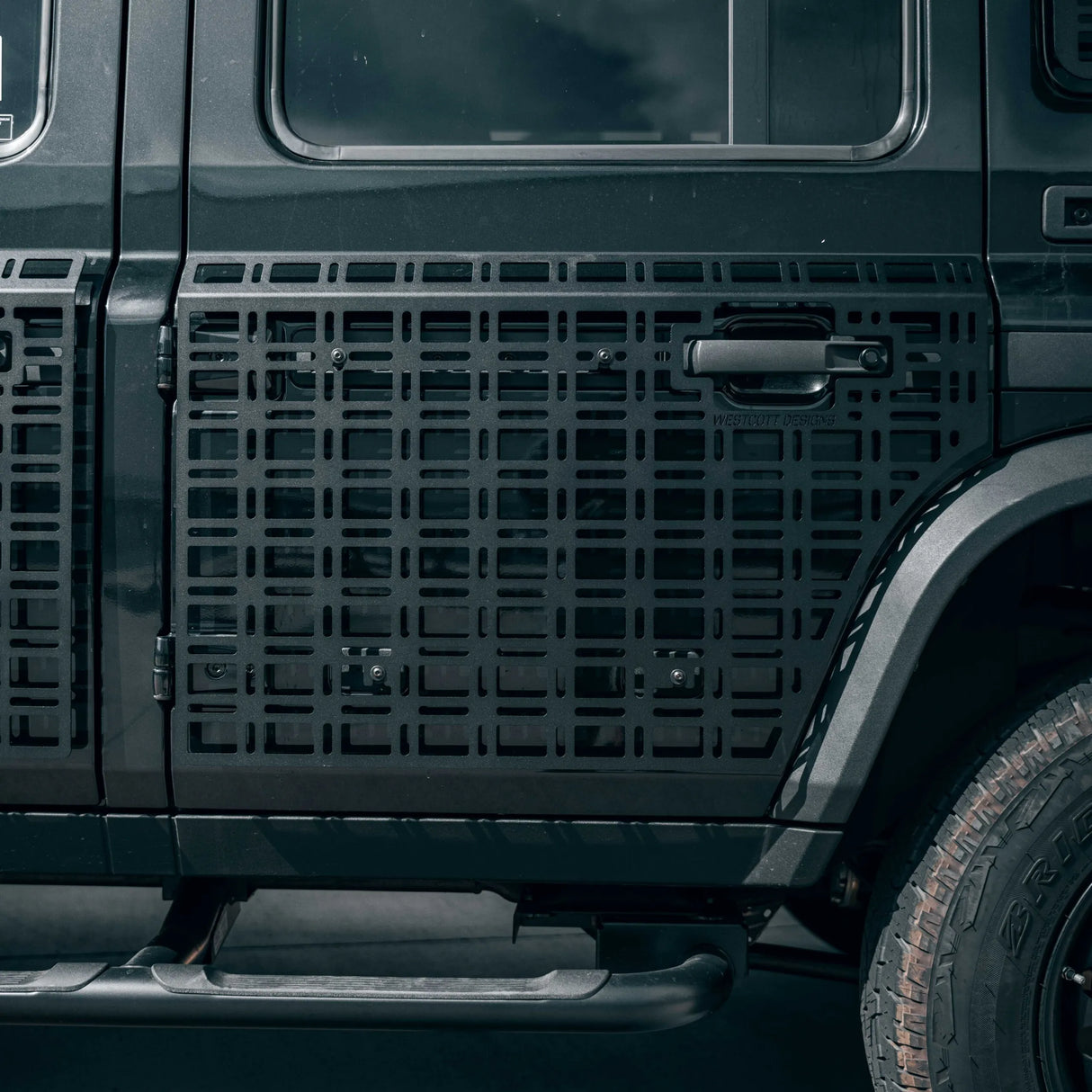 Close-up of a black vehicle side panel featuring a large black metal grid protective grille or guard over the door and side area, with part of the tire and dark body visible.