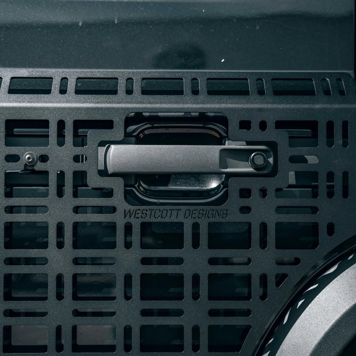 Close-up of a dark metallic handle with the brand name WESTCOTT DESIGNS embossed below it, mounted on a perforated metal panel.