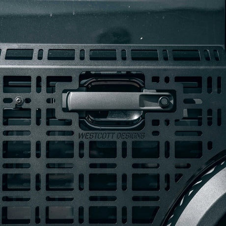 Close-up of a dark metallic handle with the brand name WESTCOTT DESIGNS embossed below it, mounted on a perforated metal panel.