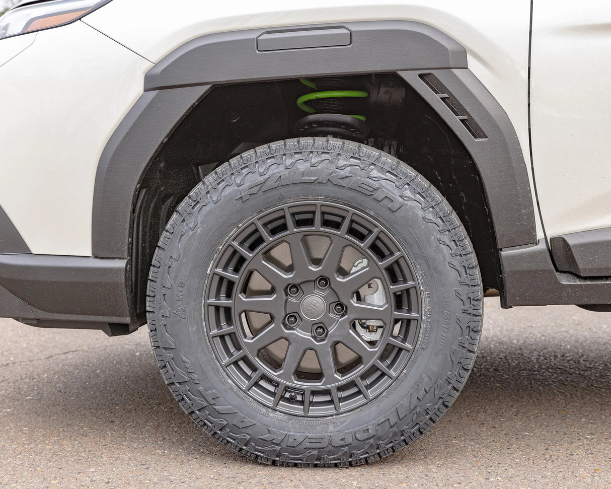 Close-up of a black off-road tire with a multi-spoke dark gray wheel, mounted on a white vehicle with black fender flares.