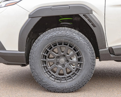 Close-up of a black off-road tire with a multi-spoke dark gray wheel, mounted on a white vehicle with black fender flares.