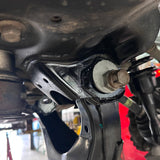 Close-up of a black metal vehicle suspension control arm bolt and bushing assembly, showing a white gasket and a rusty bolt head, photographed in a workshop setting.