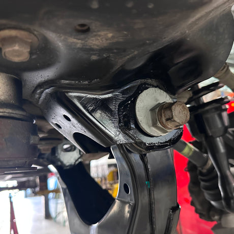 Close-up of a black metal vehicle suspension control arm bolt and bushing assembly, showing a white gasket and a rusty bolt head, photographed in a workshop setting.