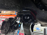 Close-up of a black metal automotive suspension or chassis component mounted underneath a vehicle, with other mechanical parts visible in a workshop setting.