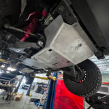 A silver metal underbody shield installed on the side of a vehicle, photographed from underneath in an auto repair shop. The vehicle has a red coilover, black tires, and other suspension components visible.