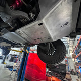 A custom brushed aluminum underbody shield installed on a vehicle, photographed from underneath in an auto repair shop. The shield features pre-drilled mounting holes and a small black logo on the right side, with a red vehicle panel and blue hydraulic lift visible in the background.