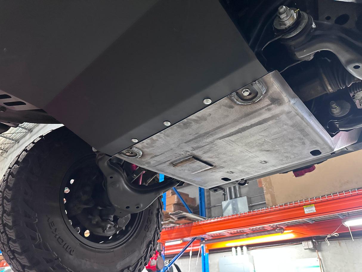 A black metal underbody shield and skid plate installed on the underside of a vehicle, shown in a garage setting with a tire and workshop equipment visible.