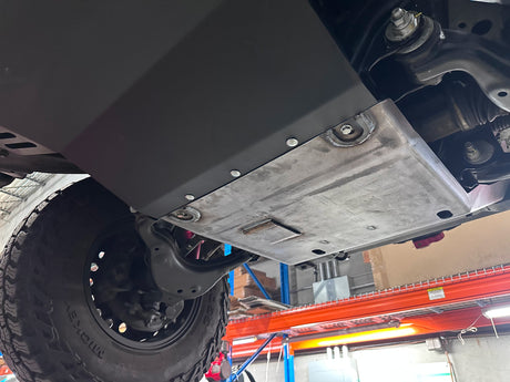 A black metal underbody shield and skid plate installed on the underside of a vehicle, shown in a garage setting with a tire and workshop equipment visible.