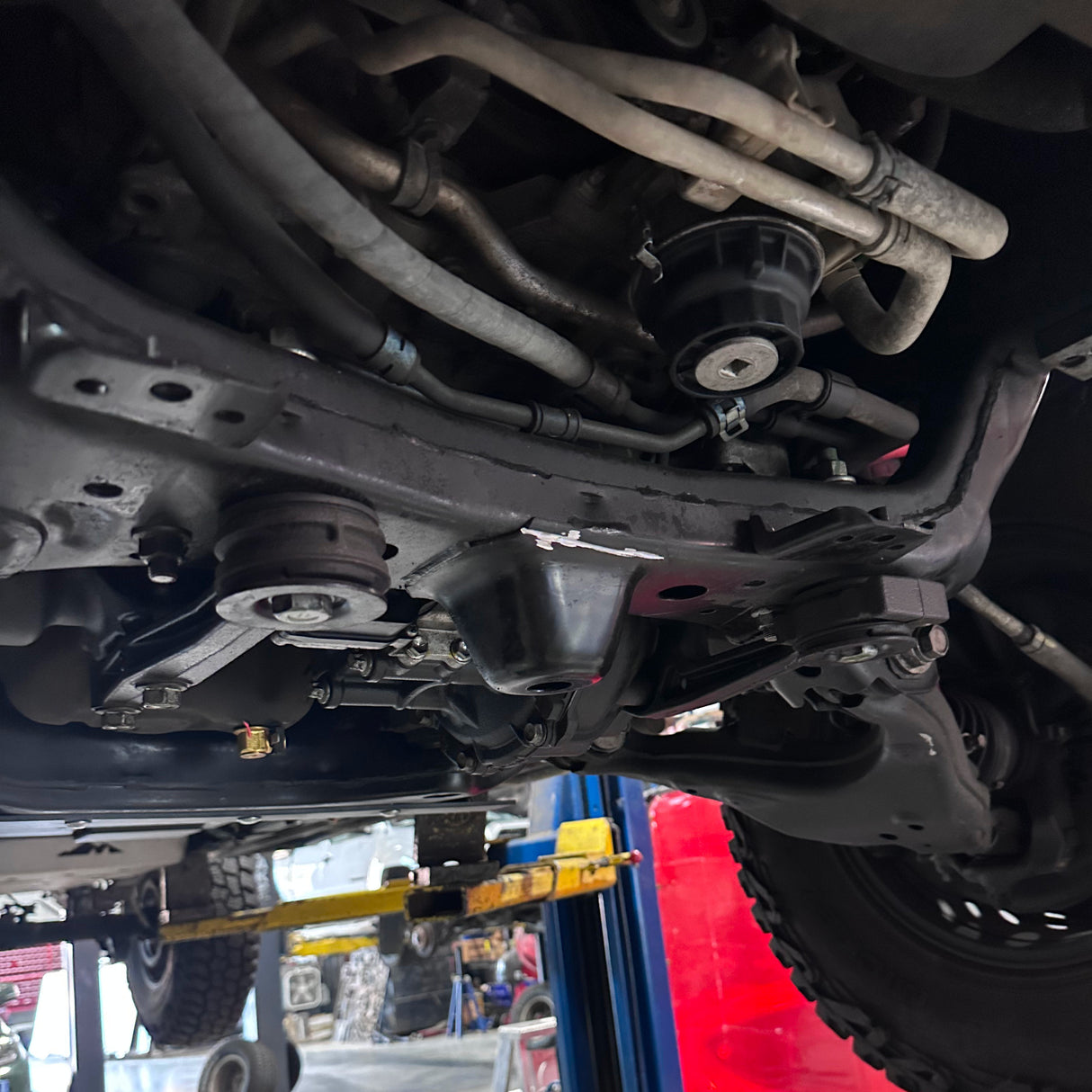 Undercarriage view of a vehicle showing suspension components, control arms, and a black oil pan, photographed in an auto repair shop with a lift visible.