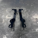 A pair of black metal mounting brackets with pre-drilled holes, displayed on a scratched gray metal surface.
