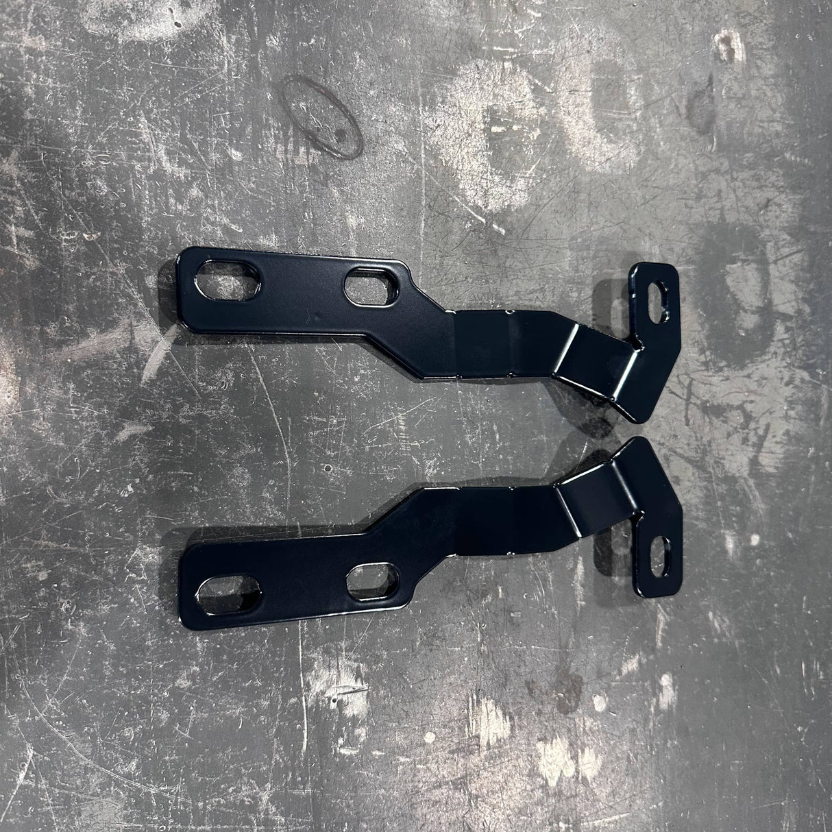 Two black metal mounting brackets with pre-drilled screw holes, laid flat on a scratched gray metal surface.