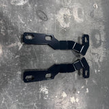 Two black metal mounting brackets with pre-drilled screw holes, laid flat on a scratched gray metal surface.