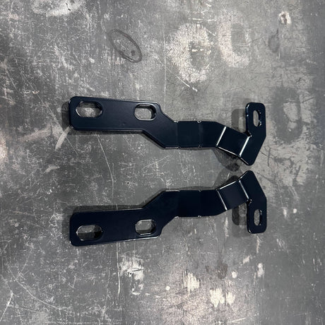 Two black metal mounting brackets with pre-drilled screw holes, laid flat on a scratched gray metal surface.