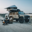 Two people sitting by a campfire in front of a large SUV with a rooftop tent in a desert setting.