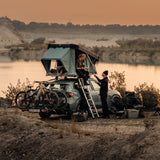 Two people setting up a rooftop tent on a car by a lake.