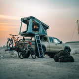 Teal rooftop tent attached to a truck with bicycles on a sandy landscape at sunset.