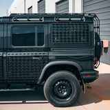 A black off-road vehicle with custom black metal grille side panels and a roof rack, parked outdoors in front of a gray building with garage doors.